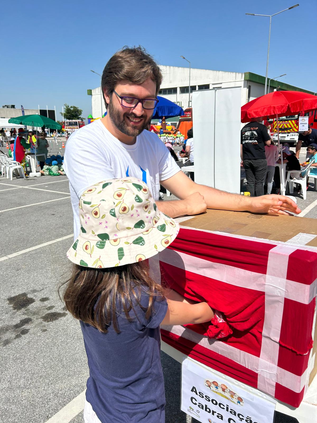 Um homem sorridente a interagir com uma criança numa banca ao ar livre, num evento em dia de sol. Em primeiro plano, à esquerda, há um homem adulto de pele clara, cabelo castanho curto e barba, usando óculos de armação escura e uma t-shirt branca. Ele está inclinado para a frente, com os braços apoiados sobre uma estrutura tipo mesa/balcão. Em frente dele, de costas para a câmara, está uma criança com cabelo castanho comprido, t-shirt azul escura e um chapéu bege com padrão de abacates. A criança parece estar a mexer ou a alcançar algo numa caixa tapada, apenas com dois buracos para colocar as mãos, sugerindo que é um jogo para adivinhar objetos dentro da caixa. A caixa está coberta com tecido vermelho. Na parte de baixo há um cartaz com texto; dá para ler “Associação” e, na linha seguinte, “Cabra Cega”. Ao fundo, vê-se um recinto amplo tipo parque de estacionamento com várias pessoas, mesas e cadeiras de plástico, chapéus de sol coloridos (azul, verde e vermelho), e um edifício grande e claro. O céu está azul limpo, indicando um dia muito ensolarado.